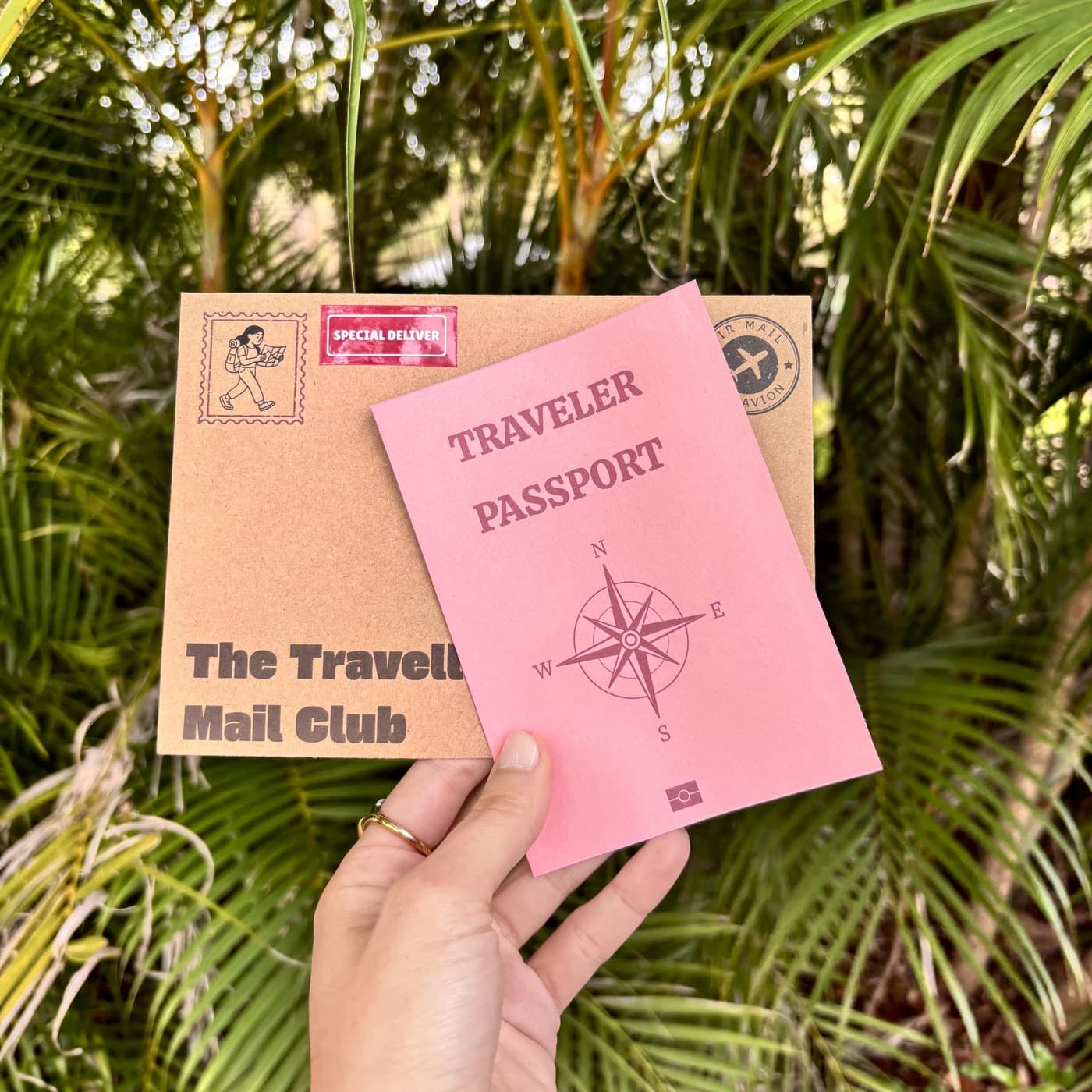 A hand holding the Traveller's Mail Club envelope and pink traveler passport in front of palm leaves